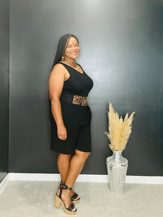 Woman in a black romperstanding next to a vase with dried plants against a black wall.