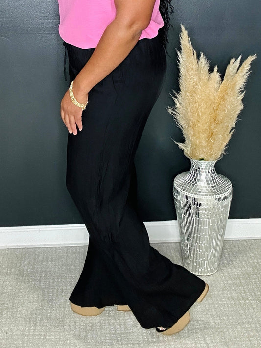 Person wearing black wide-leg pants with a pink top, standing next to a decorative vase with pampas grass against a dark wall.