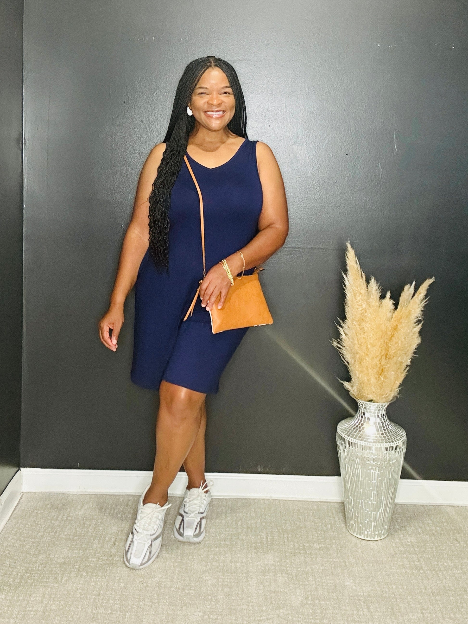 Woman in a blue romper standing next to a vase with pampas grass against a black wall.