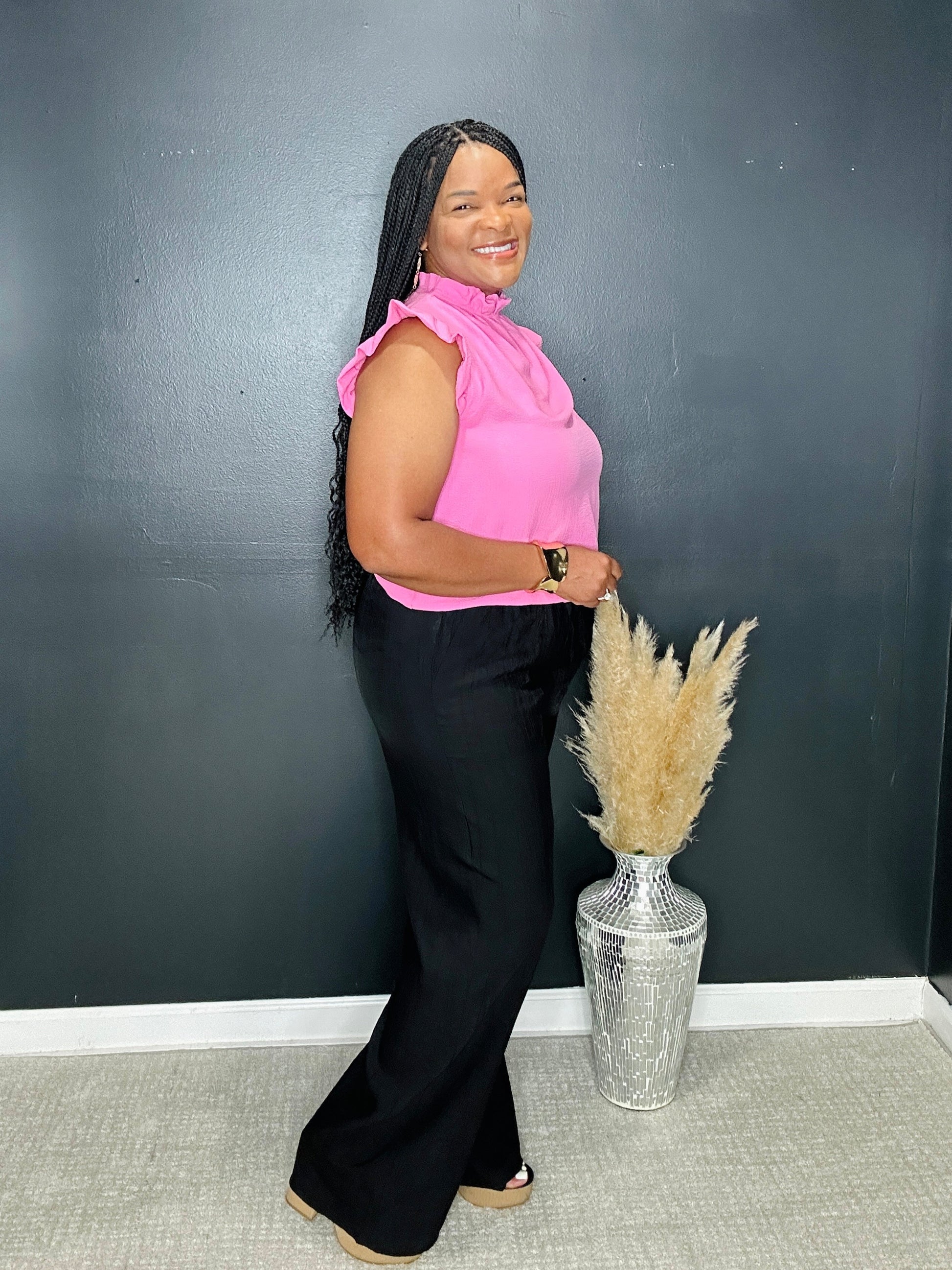 Woman in a pink top and black pants standing next to a vase with pampas grass against a dark wall.